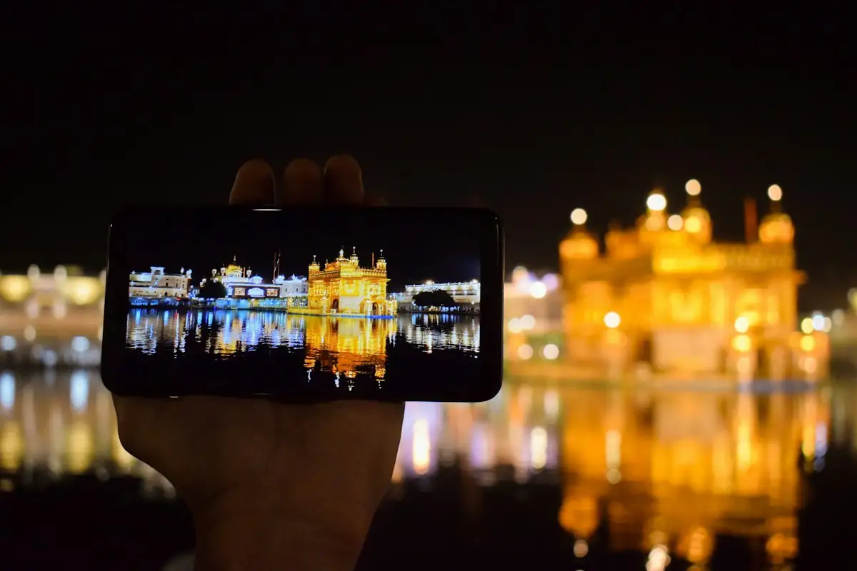 A hand holding up a smartphone that shows the Golden Temple in Amritsar, lit up at night behind it.