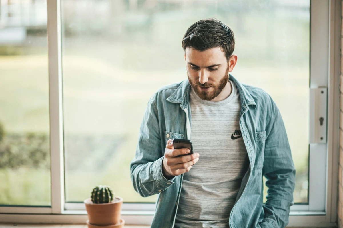 A man by a sunny window looking down at the smartphone in his hand.